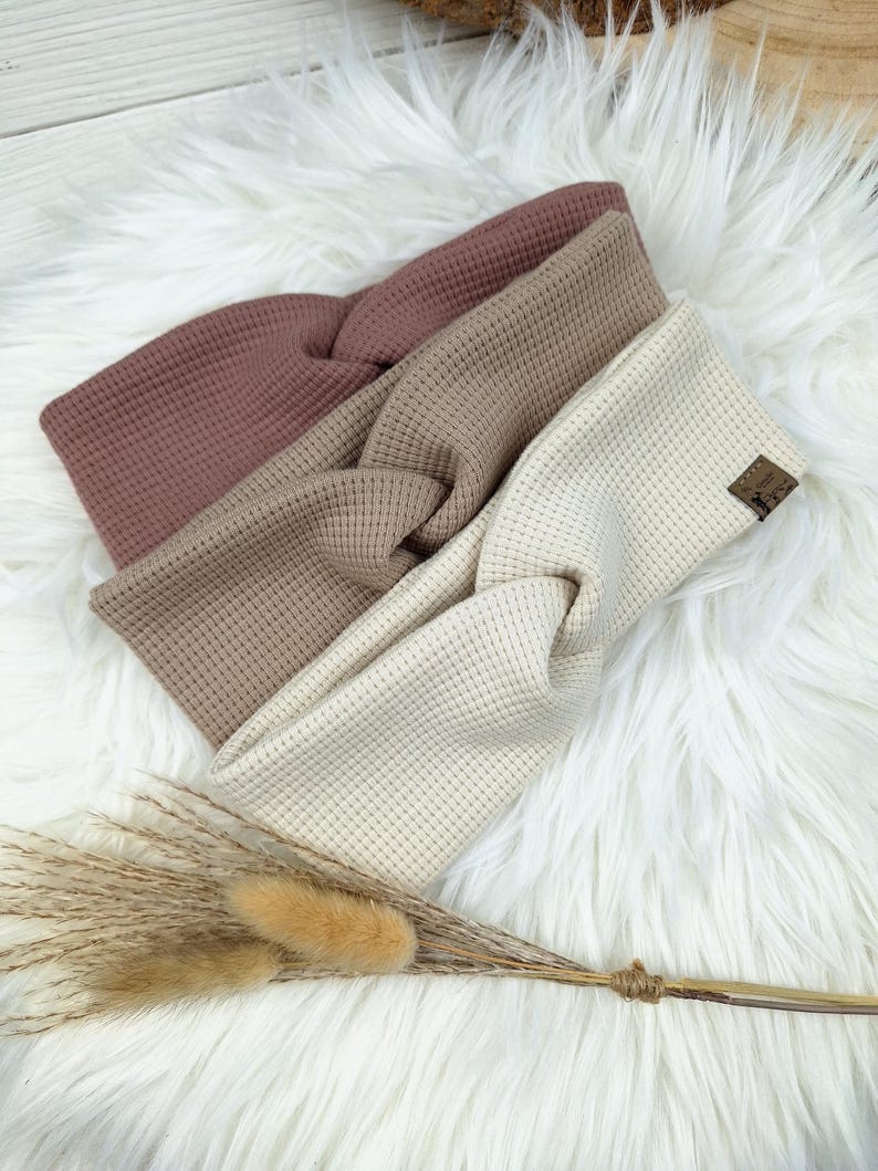 May include: Three waffle-knit headbands in shades of mauve, tan, and cream, arranged on a fluffy white surface. The headbands have a twisted knot design. A small brown label is visible on the cream headband. Dried grasses are in the foreground.