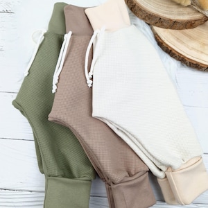 Puede incluir: Tres pares de pantalones de punto de waffle en verde, marrón y crema. Los pantalones tienen cintura con cordón y piernas con puños.