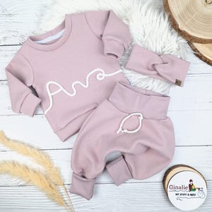 May include: A three-piece baby outfit in a soft pink hue, featuring a long-sleeved top with the name "Ana" in white script, matching pants, and a headband. The outfit is made of a textured fabric and is displayed on a white surface.