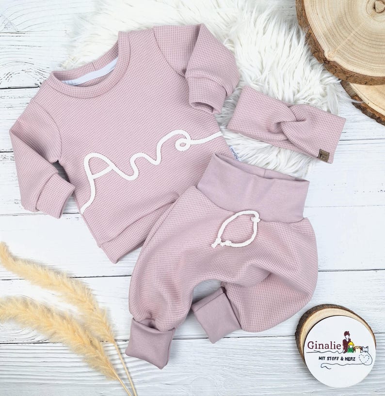 May include: A three-piece baby outfit in a soft pink hue, featuring a long-sleeved top with the name "Ana" in white script, matching pants, and a headband. The outfit is made of a textured fabric and is displayed on a white surface.