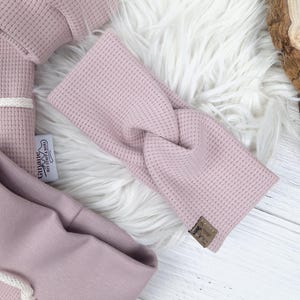 May include: A light pink waffle-knit headband with a twisted design, lying on a white faux fur surface. The headband has a small brown tag. Other matching light pink waffle-knit clothing items are also visible.