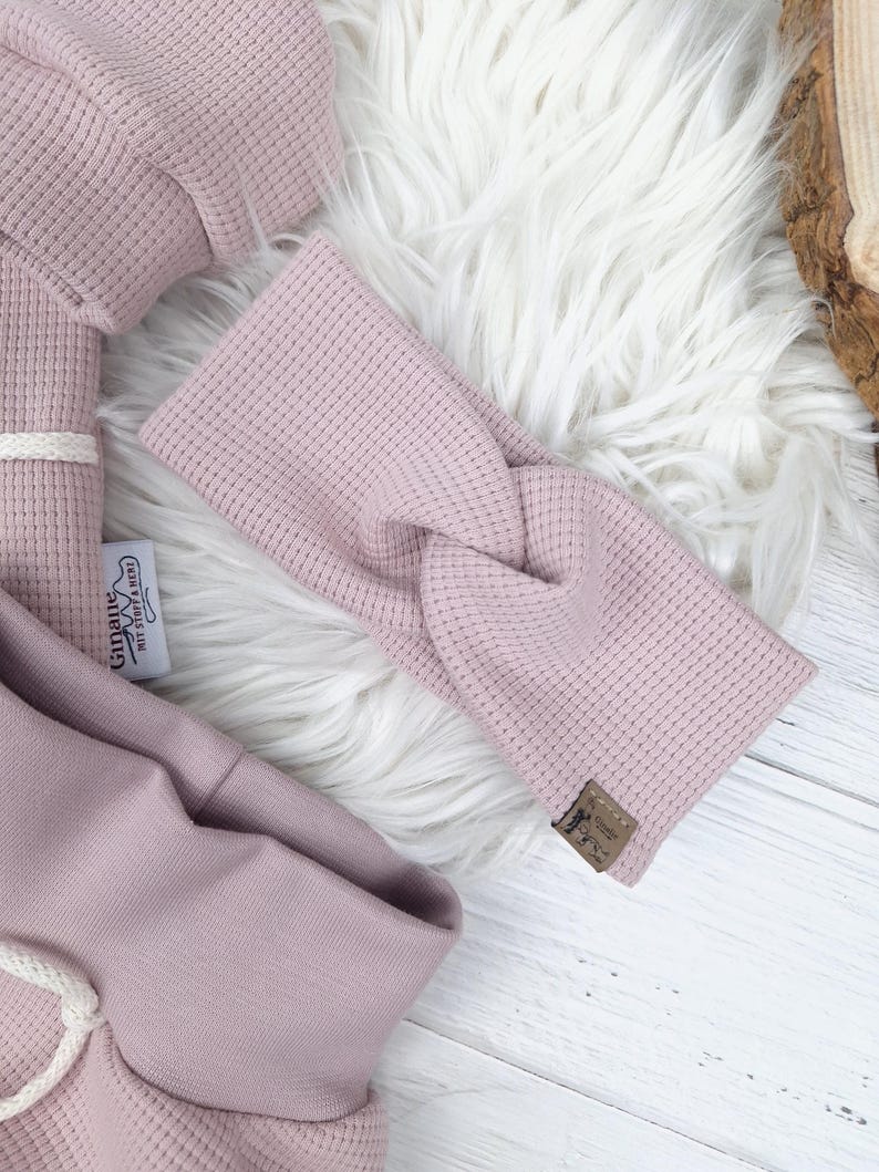 May include: A light pink waffle-knit headband with a twisted design, lying on a white faux fur surface. The headband has a small brown tag. Other matching light pink waffle-knit clothing items are also visible.