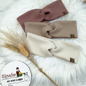 May include: Three waffle-knit headbands in dusty rose, tan, and cream, with a twisted knot design. Each headband has a small, rectangular label. The headbands are displayed on a fluffy white surface, with a decorative sprig and a round sign that reads "Ginalie".