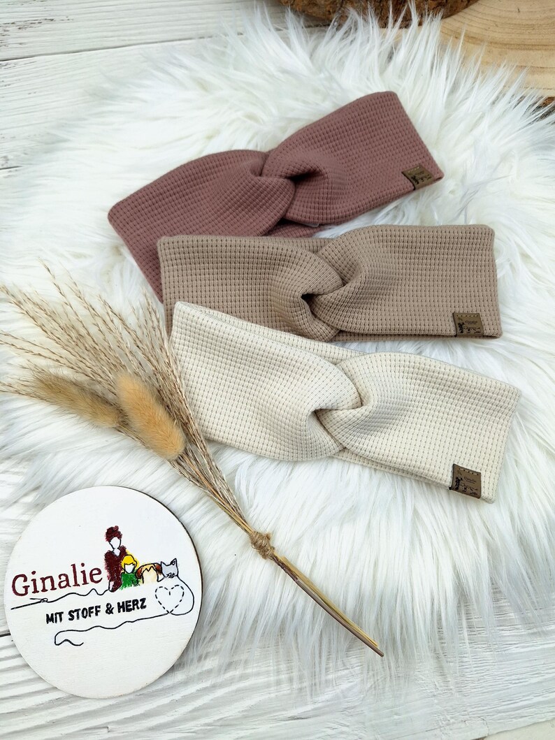May include: Three waffle-knit headbands in dusty rose, tan, and cream, with a twisted knot design. Each headband has a small, rectangular label. The headbands are displayed on a fluffy white surface, with a decorative sprig and a round sign that reads "Ginalie".