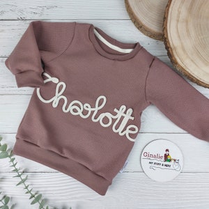 Könnte beinhalten: Ein langärmliges Sweatshirt aus mauvefarbenem Waffelpiqué mit dem Namen "Charlotte", der in weißer Farbe gestickt ist. Das Sweatshirt hat ein weißes Etikett mit dem Text "Ginalie mit Stoff & Herz".