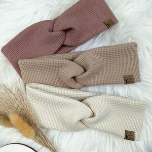 May include: Three twisted headbands in shades of mauve, tan, and cream, arranged on a fluffy white surface. Each headband features a textured waffle knit design and a small, rectangular label. The headbands are a stylish accessory.
