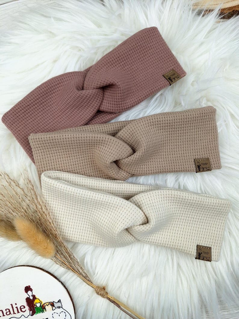 May include: Three twisted headbands in shades of mauve, tan, and cream, arranged on a fluffy white surface. Each headband features a textured waffle knit design and a small, rectangular label. The headbands are a stylish accessory.