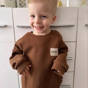 Può includere: Un bambino piccolo che indossa una maglietta a maniche lunghe in maglia a nido d'ape marrone con una toppa che dice "MUT AUS BRUCH".