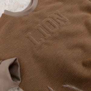 Op de afbeelding: Een bruine corduroy sweatshirt met het woord "LION" geborduurd over de borst. De sweatshirt heeft een contrasterende beige halslijn en manchetten. De stof heeft een geribbelde textuur. Het kledingstuk wordt gepresenteerd op een witte, zachte ondergrond.
