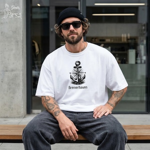 May include: A white short-sleeved t-shirt featuring a black graphic of an anchor centered over a compass rose, with the text Bremerhaven printed below the design. The shirt is worn with dark gray jeans, a black beanie, and black sunglasses.
