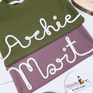 Può includere: Due maglie a nido d'ape con nomi ricamati in bianco, "Archie" e "Mort", su una superficie di legno bianco. Le maglie sono verdi e malva. Una piccola etichetta rotonda con il testo "Ginalie mit Stoff & Herz" è visibile in basso a destra.