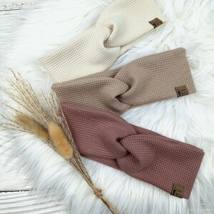 May include: Three waffle-knit headbands in cream, taupe, and mauve, each with a knotted design. The headbands are displayed on a white faux fur surface, with dried ornamental grasses to the left. Each headband has a small brown label.
