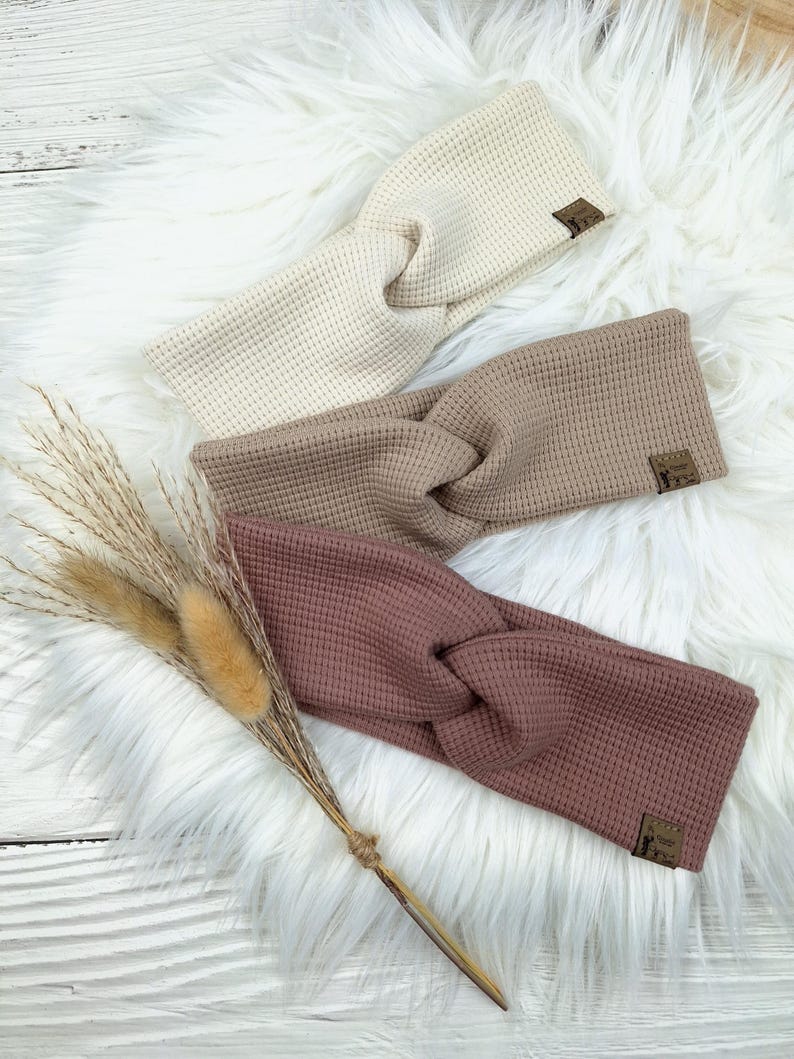 May include: Three waffle-knit headbands in cream, taupe, and mauve, each with a knotted design. The headbands are displayed on a white faux fur surface, with dried ornamental grasses to the left. Each headband has a small brown label.