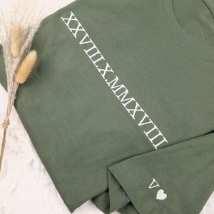 May include: Olive green t-shirt with white Roman numerals running down the left sleeve. The sleeve cuff features the letter "V" and a small white heart. The shirt is made of a soft material.