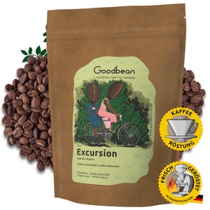 Op de afbeelding: Een bruine papieren zak met een label waarop staat "Goodbean A Tasteful Trip to Uganda". Het label toont een illustratie van twee mensen die op een fiets door een koffieplantage rijden. De tekst "The Excursion Uganda | Bugisu" staat ook op het label. De zak is gevuld met geroosterde koffiebonen.