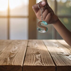May include: A resin keychain in the shape of the letter "S" is held up. The keychain has a silver chain and ring. The letter is a mix of brown, white, and turquoise colors. The background is blurred, with a wooden table in the foreground.