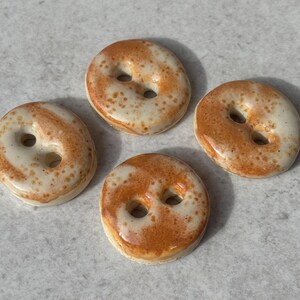 May include: Four round ceramic buttons featuring a glossy off-white base with speckled brown and tan glaze patterns. Each button has two central holes for attachment, a smooth rounded edge, and a reflective finish across the surface.