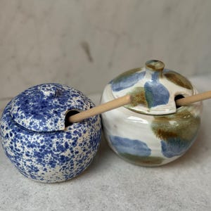 May include: Two round ceramic jars with lids and wooden spoons. The left jar features a blue and white speckled pattern. The right jar has a white base with abstract blue and brown painted patches. Both containers have a small knob on the lid.