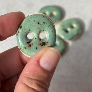 May include: A set of round, two-hole ceramic buttons featuring a glossy light green glaze with dark brown speckles and spots. The buttons have a smooth, slightly irregular surface texture and a visible off-white base material along the edges.