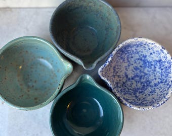 May include: A set of four ceramic bowls with pouring spouts. One bowl is dark blue-green, one is light blue with brown speckles, one is deep teal, and one is white with a dense blue speckled pattern. Each bowl features a rounded shape and a spout.