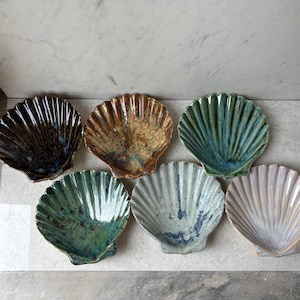 May include: A collection of six ceramic seashell-shaped dishes in various colors. The dishes feature a ribbed texture and glossy finish. Colors include dark brown, tan, green, blue, and off-white. These decorative dishes are ideal for serving small portions.