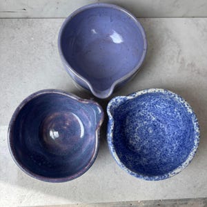 May include: Three ceramic bowls in shades of blue. Each bowl has a pouring spout. The top bowl is a solid lavender color. The bottom left bowl is a deep indigo with purple hues. The bottom right bowl is speckled blue.
