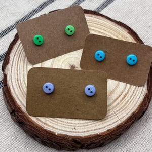 Könnte beinhalten: Drei Paar Knopf-Ohrringe in Grün, Blau und Lavendel, jeweils auf einer kleinen braunen Karte befestigt. Die Ohrringe werden auf einer Holzscheibe vor einem neutralen Stoffhintergrund präsentiert.