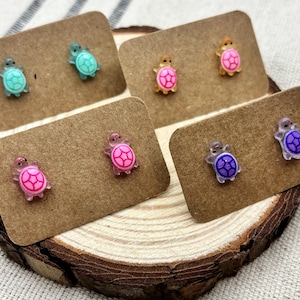 Op de afbeelding: Een verzameling kleurrijke schildpad oorknopjes, gepresenteerd op bruine oorbellenkaarten. De oorbellen hebben verschillende kleuren, waaronder turquoise, roze en paars. De oorbellen worden getoond op een houten schijf.