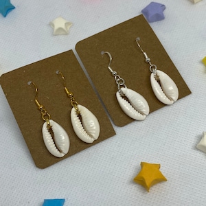 May include: Two pairs of earrings with white shell charms hanging from gold and silver chains. The earrings are displayed on brown card stock.
