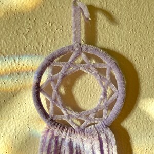 May include: A lavender dreamcatcher with a circular frame and a star-shaped web design. The dreamcatcher has a loop for hanging and soft, fringed tassels hanging below. The background is a textured, light-colored wall.