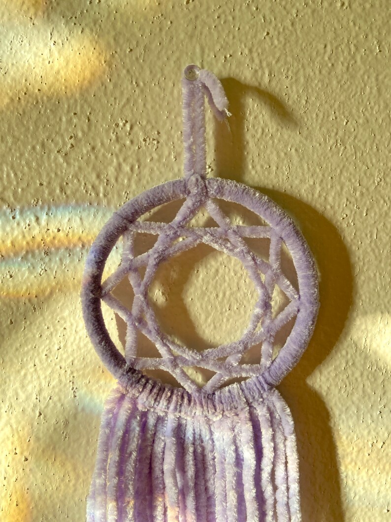May include: A lavender dreamcatcher with a circular frame and a star-shaped web design. The dreamcatcher has a loop for hanging and soft, fringed tassels hanging below. The background is a textured, light-colored wall.