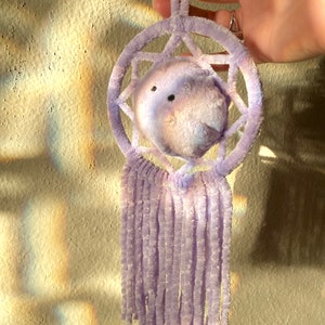 May include: A handmade dreamcatcher in shades of lavender and white. The circular frame and central sphere are made of soft, fuzzy material. The dreamcatcher features a star-like pattern within the circle and dangling fringe.