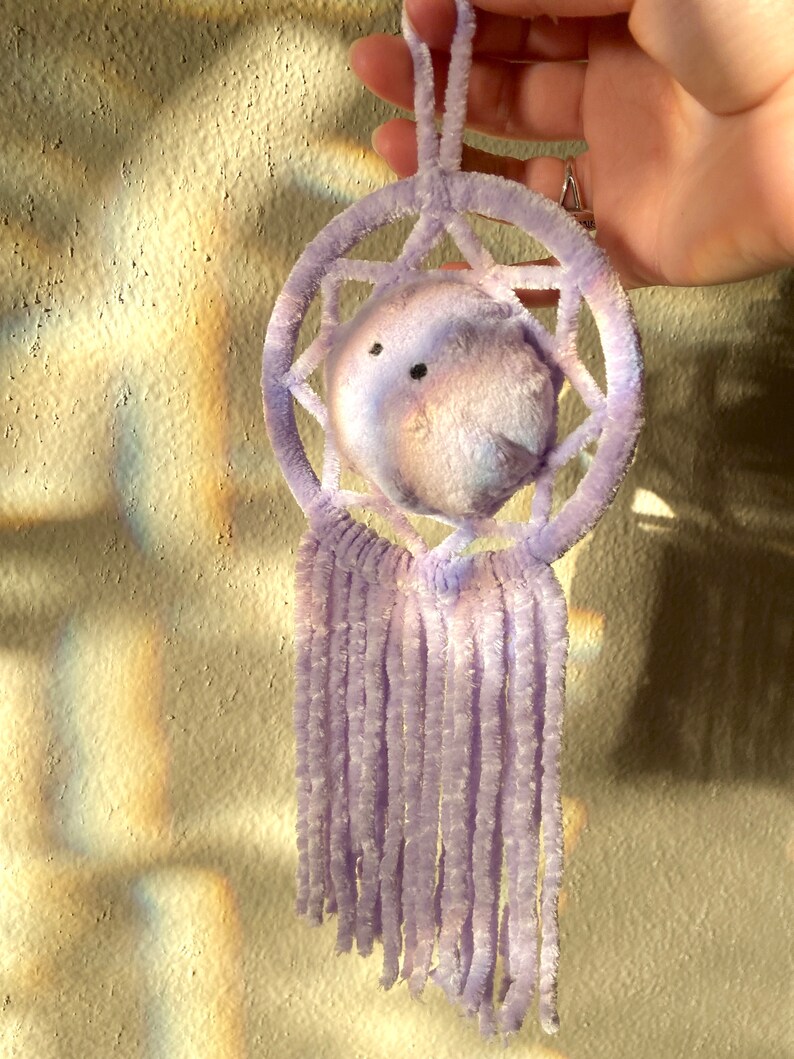 May include: A handmade dreamcatcher in shades of lavender and white. The circular frame and central sphere are made of soft, fuzzy material. The dreamcatcher features a star-like pattern within the circle and dangling fringe.