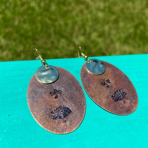 May include: A pair of oval-shaped earrings with a dragonfly and tree design. The earrings are made of brass and have a gold-tone finish.