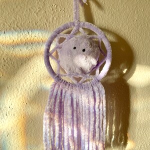 May include: A lavender dreamcatcher with a plush, round center and dangling fringe. The dreamcatcher is hanging against a textured, off-white wall, with a shadow cast to the right. The design features a circular frame and a soft, fuzzy texture.