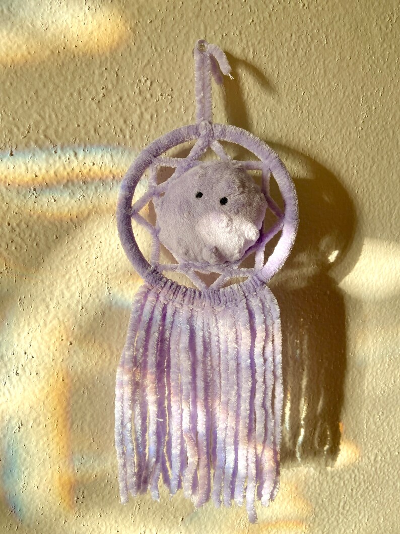 May include: A lavender dreamcatcher with a plush, round center and dangling fringe. The dreamcatcher is hanging against a textured, off-white wall, with a shadow cast to the right. The design features a circular frame and a soft, fuzzy texture.
