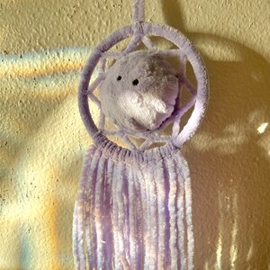 May include: A lavender dreamcatcher with a plush, round center featuring black eyes and a nose. The circular frame and hanging strands are made of soft, fuzzy material. The dreamcatcher is hanging on a textured, off-white wall.