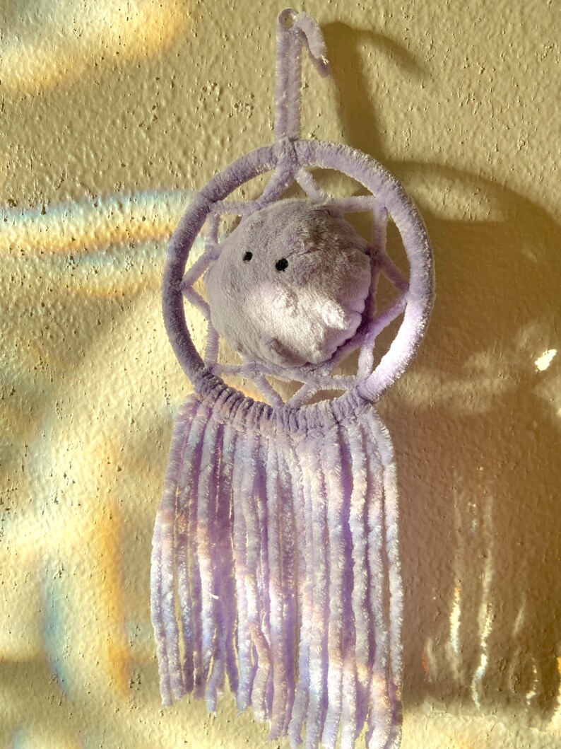 May include: A lavender dreamcatcher with a plush, round center featuring black eyes and a nose. The circular frame and hanging strands are made of soft, fuzzy material. The dreamcatcher is hanging on a textured, off-white wall.
