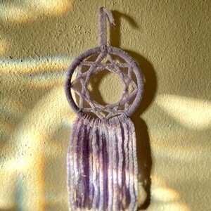 May include: A lavender dreamcatcher with a star-shaped web design and long, soft fringe. The circular frame is suspended by a loop, and the dreamcatcher hangs against a textured, light-colored wall.