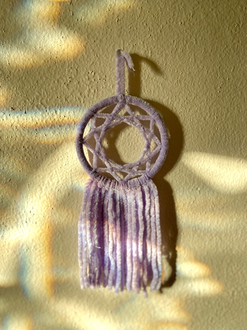 May include: A lavender dreamcatcher with a star-shaped web design and long, soft fringe. The circular frame is suspended by a loop, and the dreamcatcher hangs against a textured, light-colored wall.