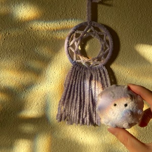 May include: A lavender dreamcatcher with a star-shaped design and fringe hangs on a textured wall. A small, round, lavender plush toy with black eyes is held in a hand, next to the dreamcatcher.