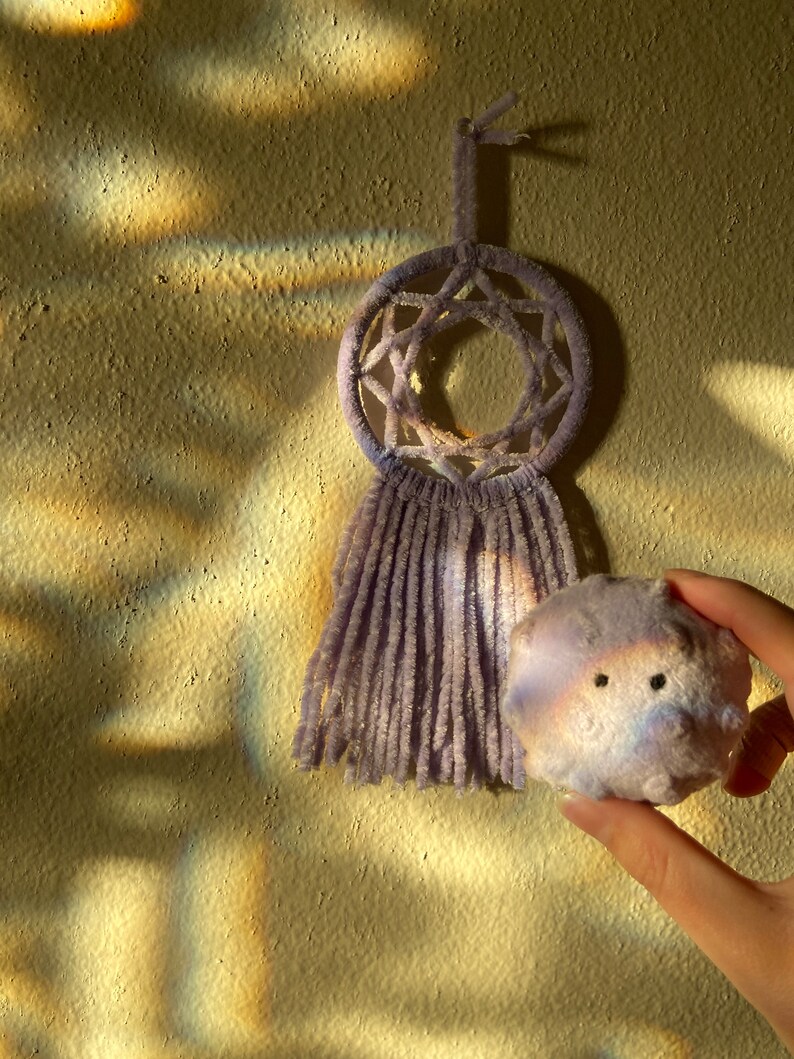 May include: A lavender dreamcatcher with a star-shaped design and fringe hangs on a textured wall. A small, round, lavender plush toy with black eyes is held in a hand, next to the dreamcatcher.