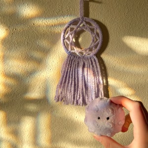 May include: A lavender dreamcatcher with a star-shaped web design and fringe hangs on a textured wall. A small, round, lavender plush toy with two black eyes is held in a hand, positioned near the dreamcatcher.