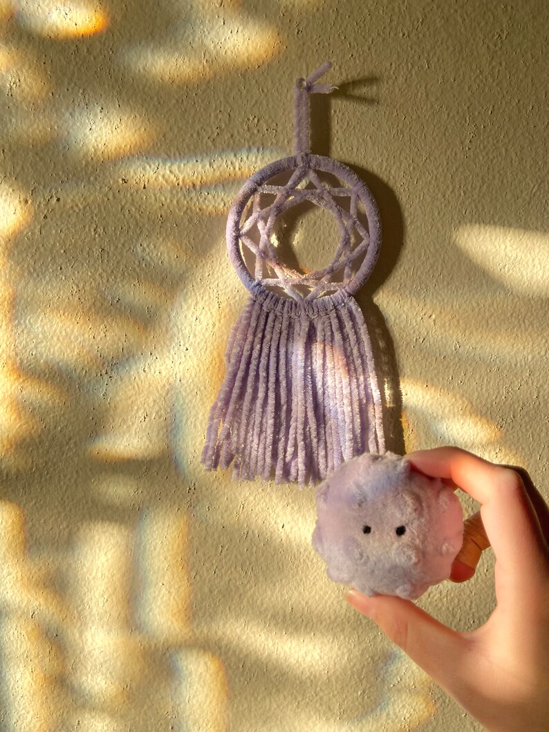 May include: A lavender dreamcatcher with a star-shaped web design and fringe hangs on a textured wall. A small, round, lavender plush toy with two black eyes is held in a hand, positioned near the dreamcatcher.