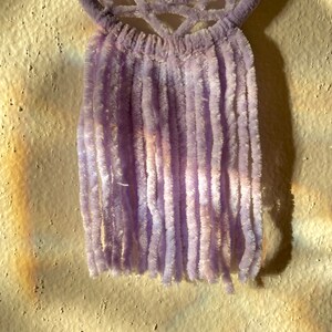 May include: A lavender-colored, handmade wall hanging. The top features a woven design, and the bottom has long, vertical strands. The soft, fuzzy texture and light purple hue create a delicate and artistic aesthetic.