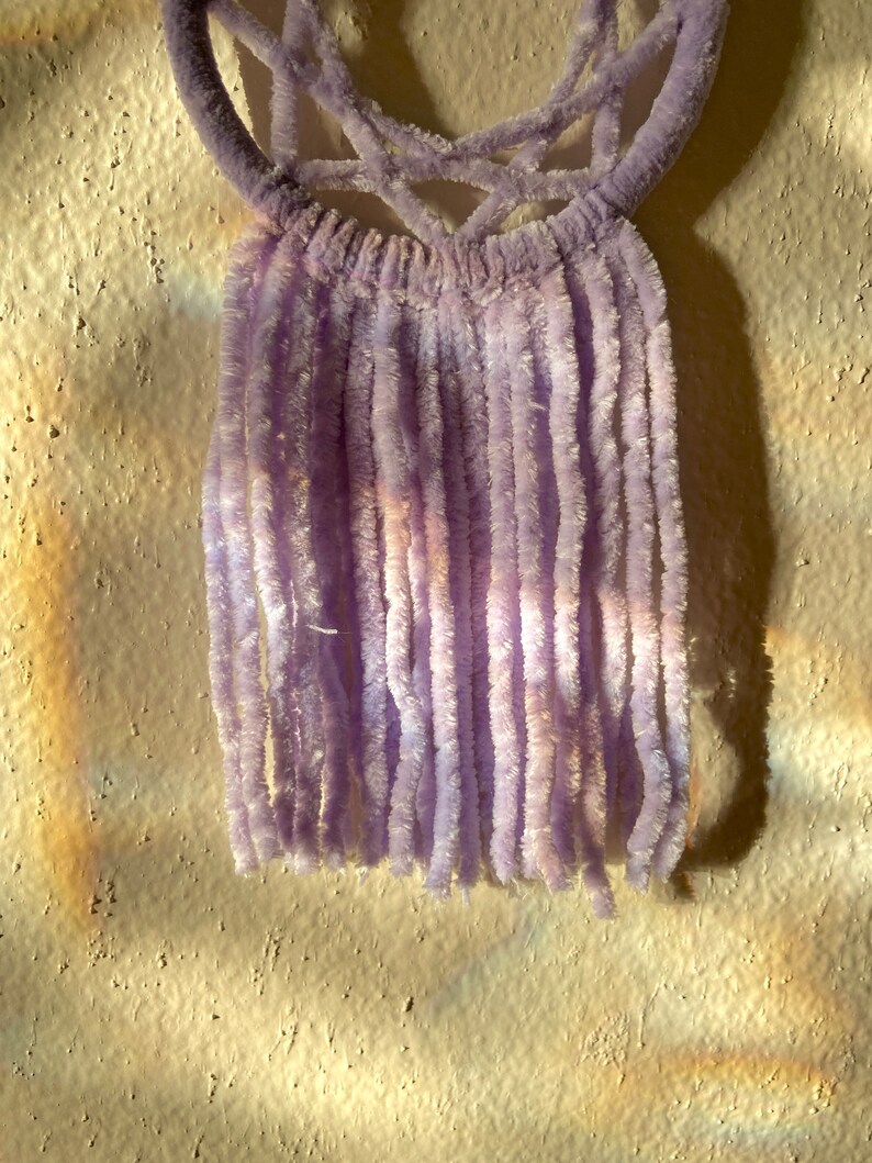 May include: A lavender-colored, handmade wall hanging. The top features a woven design, and the bottom has long, vertical strands. The soft, fuzzy texture and light purple hue create a delicate and artistic aesthetic.