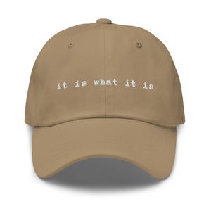 May include: A tan dad hat with the text "it is what it is" embroidered in white on the front.