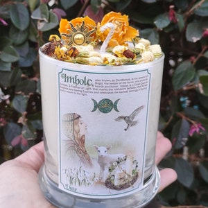 May include: A white candle with dried flowers and a sun-shaped charm on top. The candle has a label with the word "Imbolc" and illustrations of a woman, lamb, and bird. The candle is in a clear glass holder.