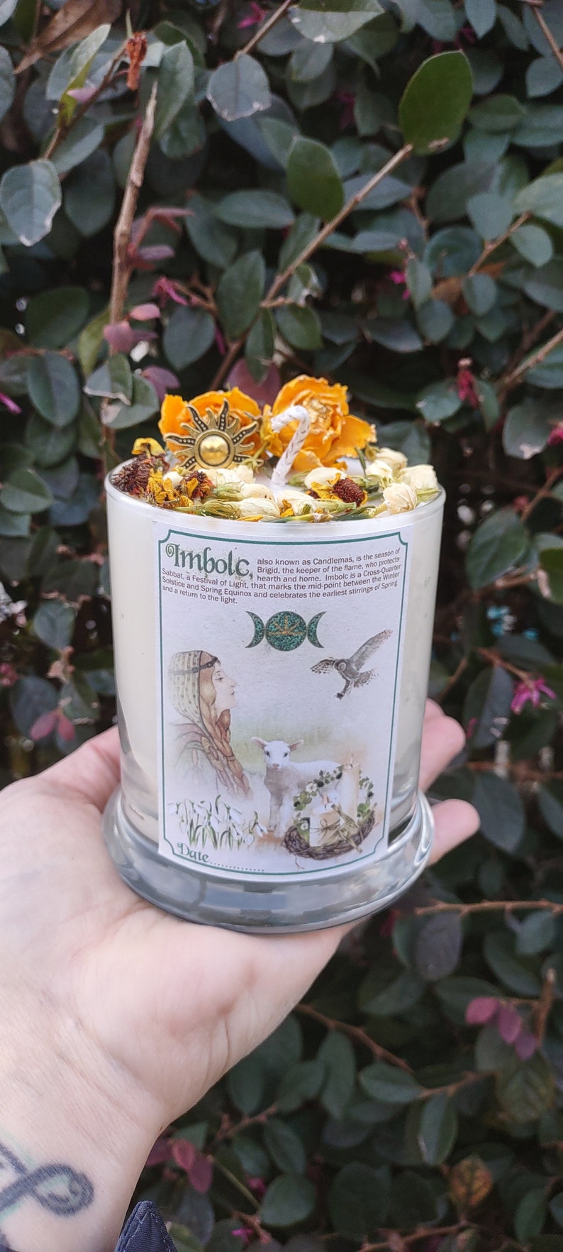 May include: A white candle with dried flowers and a sun-shaped charm on top. The candle has a label with the word "Imbolc" and illustrations of a woman, lamb, and bird. The candle is in a clear glass holder.