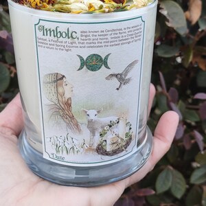 May include: A candle in a clear glass jar, decorated with dried flowers and a sun-shaped charm. The label reads "Imbolc" and features an illustration of a woman, lamb, and bird. The candle is held in a hand.
